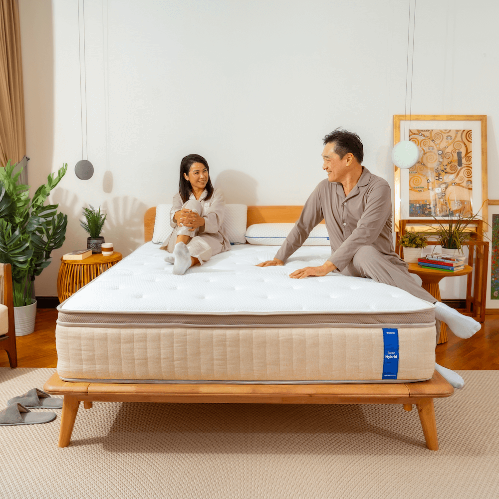 Man and woman sitting on a mattress in a bedroom setting with plants and decor.
