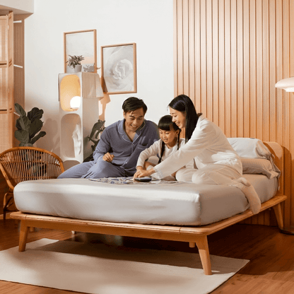 Family of three sitting on a wooden bed in a bright bedroom.