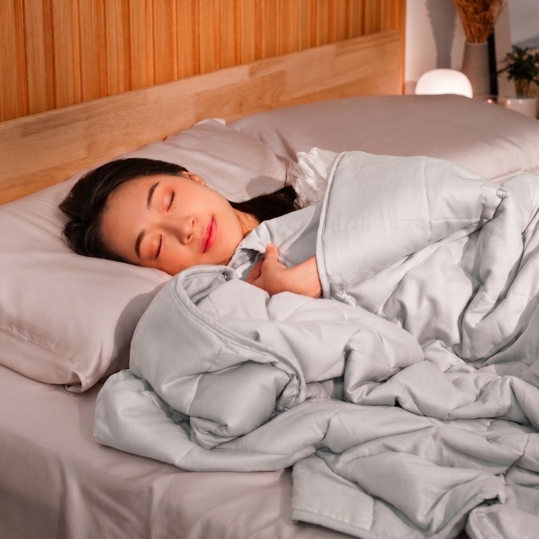 
A young woman sleeping soundly in a cozy bed with a light grey quilted comforter and matching pillows. The scene features a warm wooden headboard and soft ambient bedside lighting, creating a peaceful and comfortable atmosphere for sleep wellness and home decor 