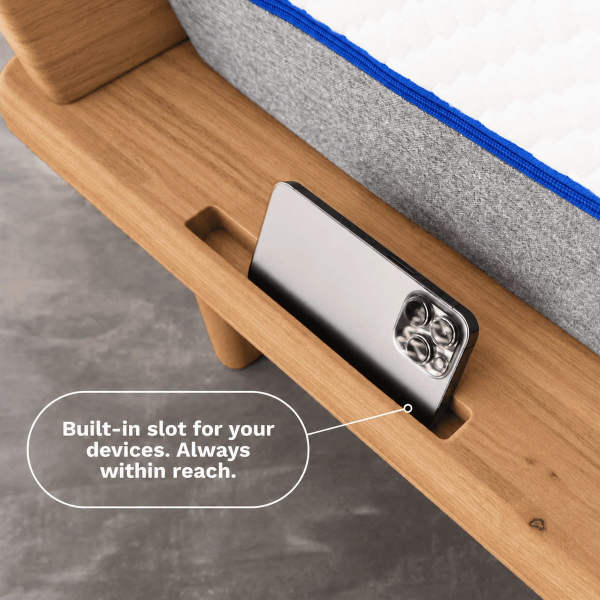 Close-up of a wooden Sonno Signature bed frame with a built-in slot holding a smartphone, with a caption reading 'Built-in slot for your devices. Always within reach.