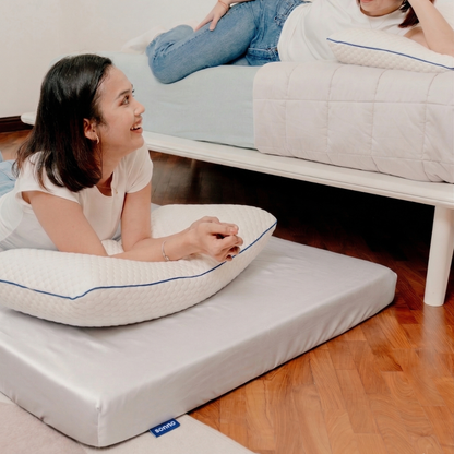 Woman lying on a Sonno Foldable mattress covered with a fitted sheet and with a pillow, another person sitting on a bed in the background.