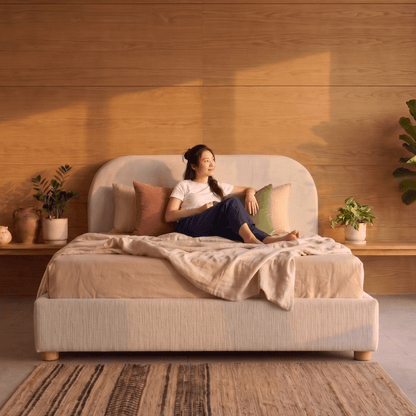 Woman sitting on a Sonno Sunset divan bed frame in a cozy bedroom with wooden walls and plants.