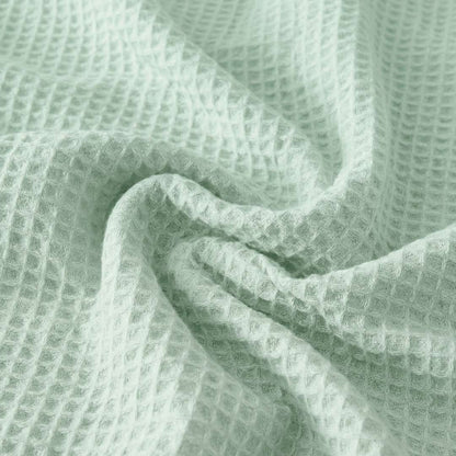Close-up of a Sonno Waffle blanket in a green color. The fabric is gathered into soft, heavy folds in the center, highlighting the depth of the "waffle" pockets. The lighting creates subtle shadows within each tiny square