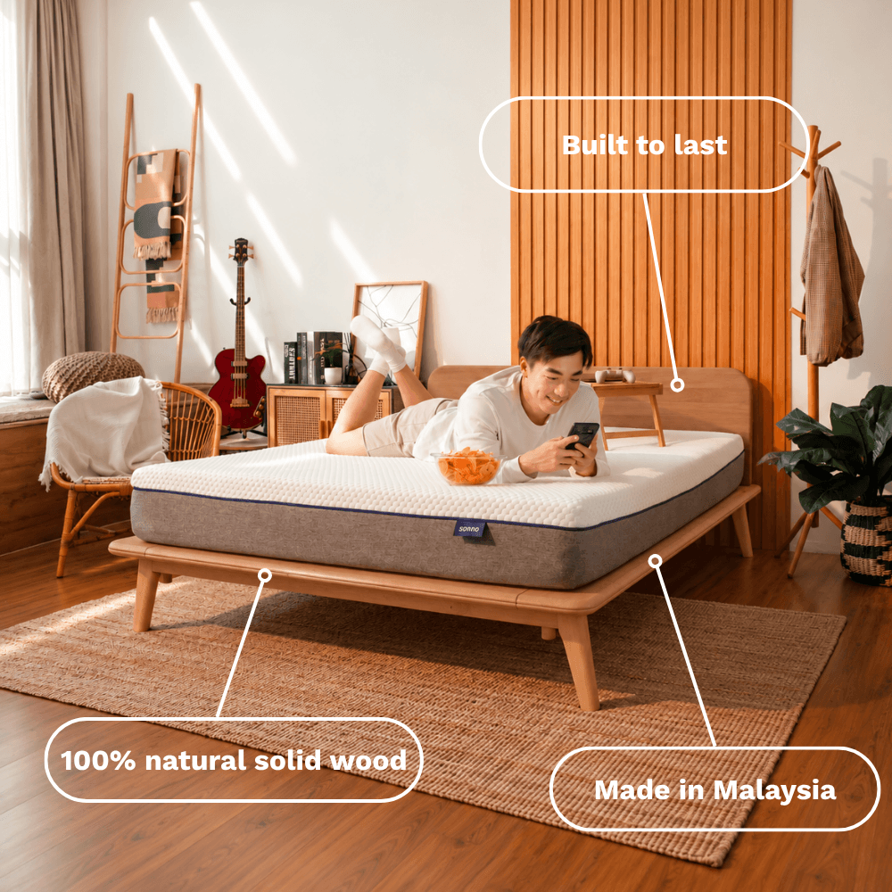 A person lies on a mattress on a Sonno Signature wooden bed frame in a styled bedroom. White callout lines with text highlight features: 'Built to last,' '100% natural solid wood,' and 'Made in Malaysia.
