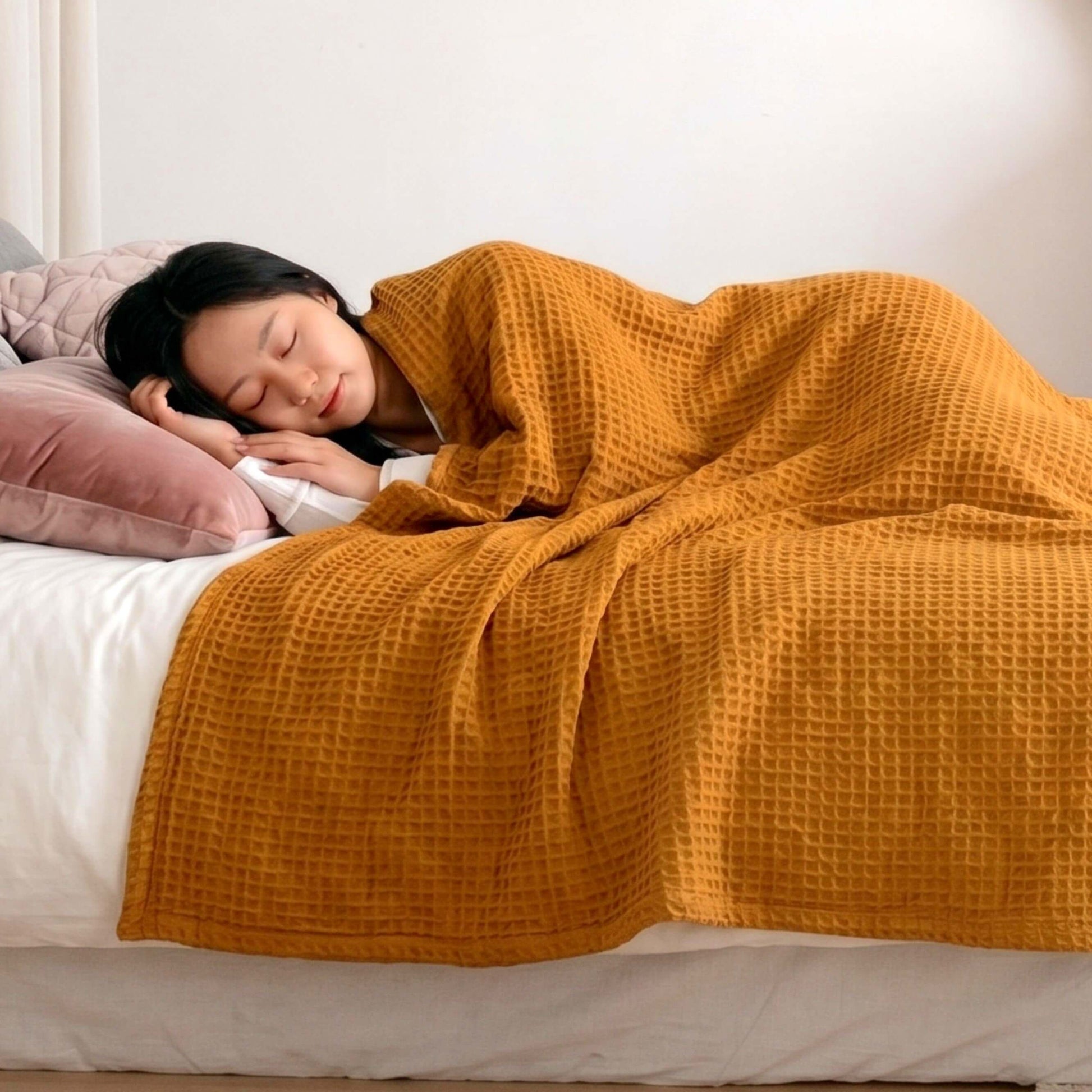 Person sleeping under a mustard yellow Sonno waffle blanket on a bed with pink pillows.