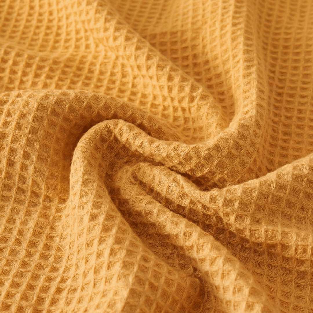 Close-up of a Sonno Waffle blanket in a warm, mustard-orange or golden-yellow hue. The fabric is gathered into soft, heavy folds in the center, highlighting the depth of the "waffle" pockets. The lighting creates subtle shadows within each tiny square