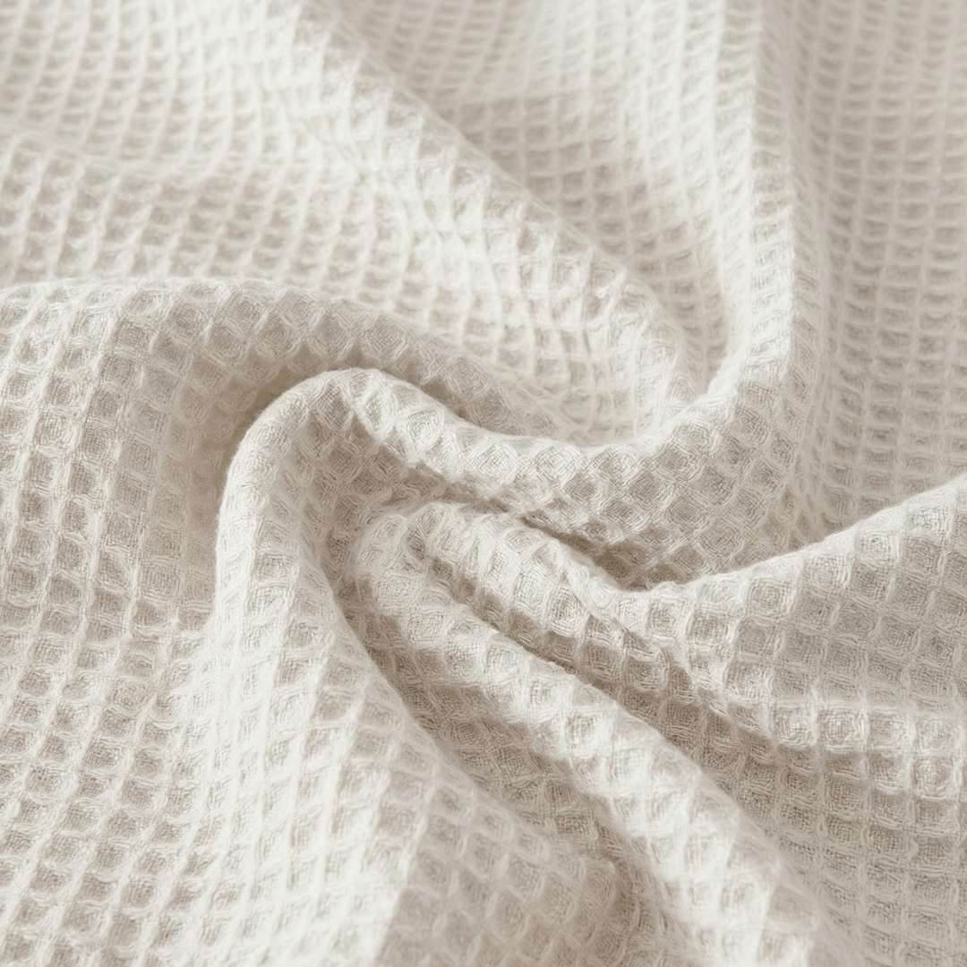 Close-up of a Sonno Waffle blanket in a white color. The fabric is gathered into soft, heavy folds in the center, highlighting the depth of the "waffle" pockets. The lighting creates subtle shadows within each tiny square.