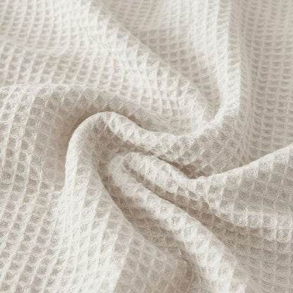 Close-up of a Sonno Waffle blanket in a white color. The fabric is gathered into soft, heavy folds in the center, highlighting the depth of the "waffle" pockets. The lighting creates subtle shadows within each tiny square.