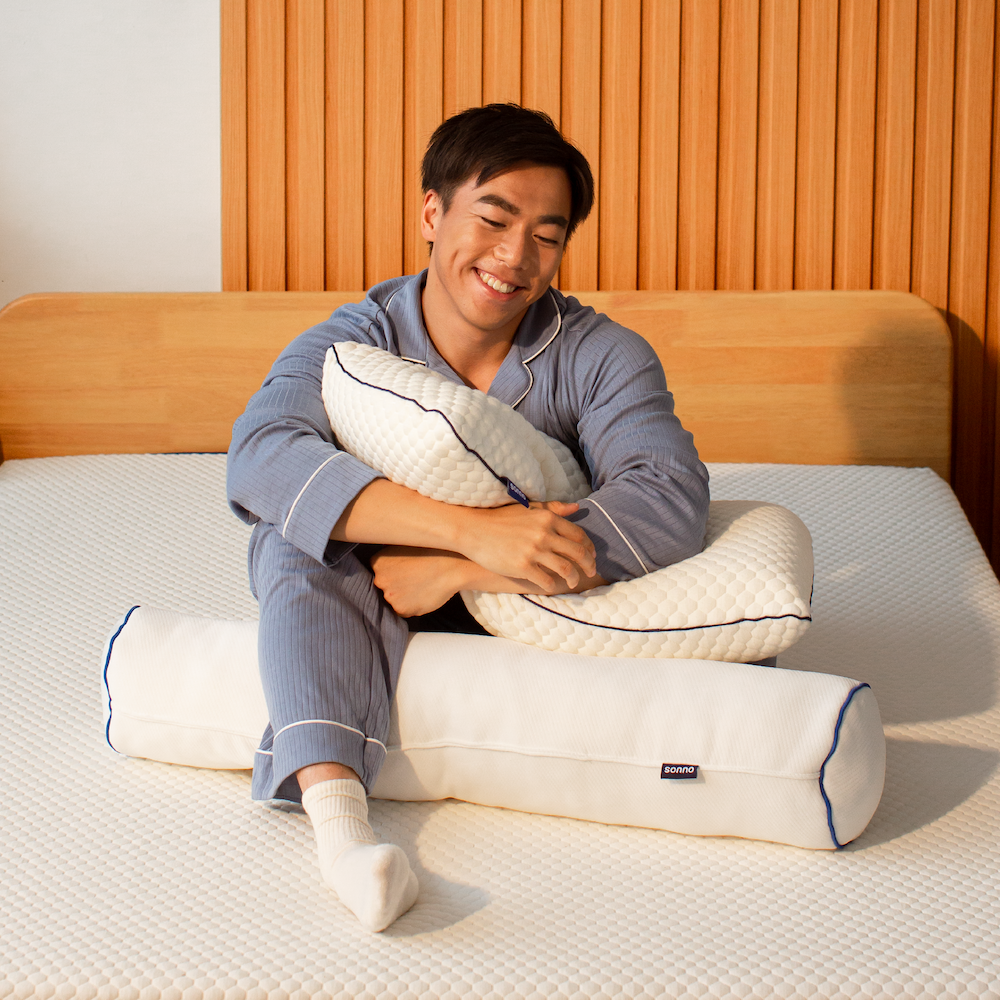 A person sitting on a bed hugging a white Sonno bolster pillow with a blue stripe.