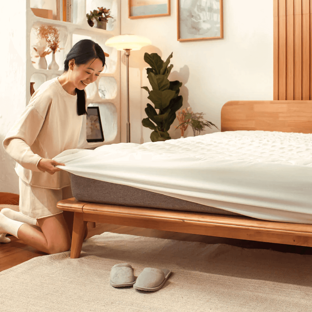 A person is fitting a white mattress protector on a bed with a wooden bed frame. The room is well-lit with natural light and has decorative plants and a shelf with framed pictures.