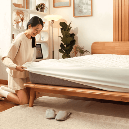 A person is fitting a white mattress protector on a bed with a wooden bed frame. The room is well-lit with natural light and has decorative plants and a shelf with framed pictures.