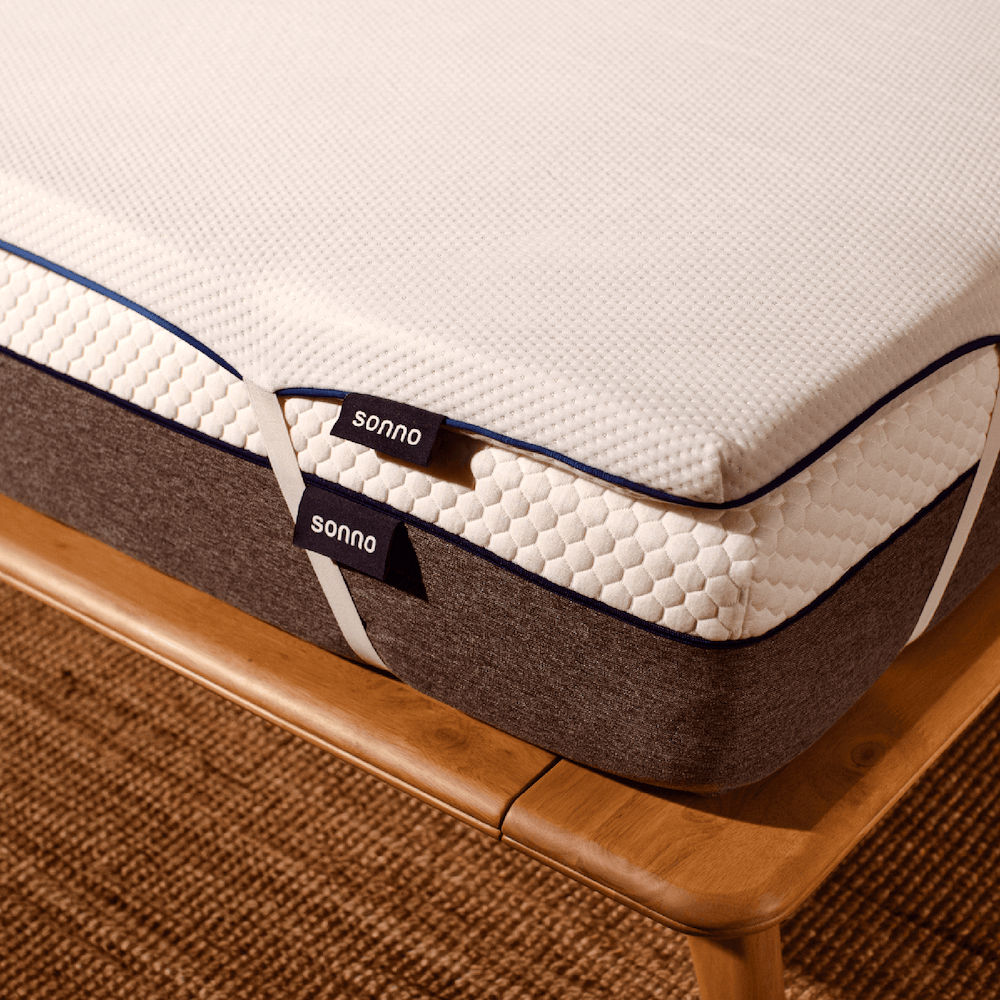 A mattress topper by Sonno, featuring a beige and gray color scheme, placed on a mattress on a wooden bed frame.