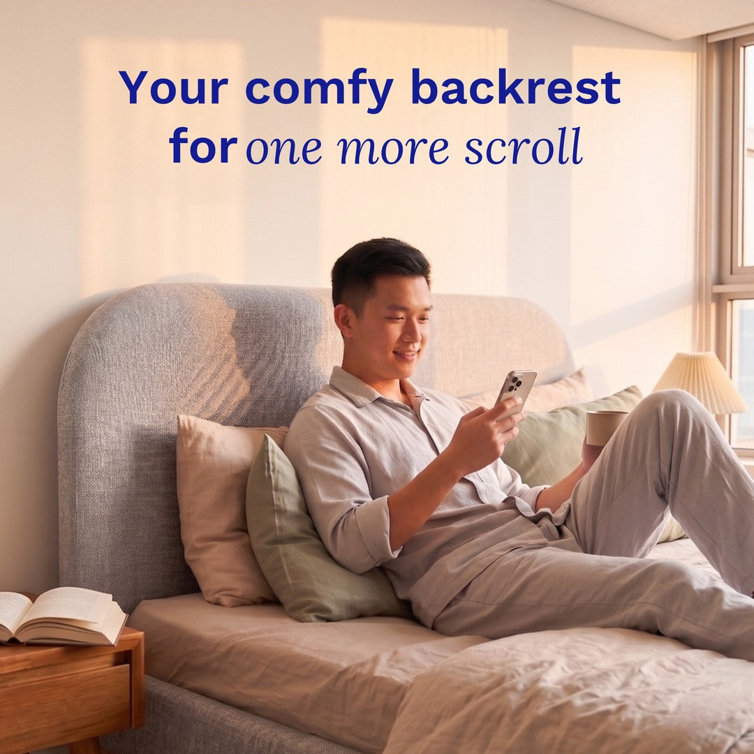 Man sitting on a Sonno Sunset Bed Frame using a smartphone with text 'Your comfy backrest for one more scroll' in a bright room.
