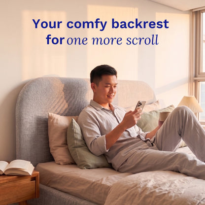 Man sitting on a Sonno Sunset Bed Frame using a smartphone with text 'Your comfy backrest for one more scroll' in a bright room.